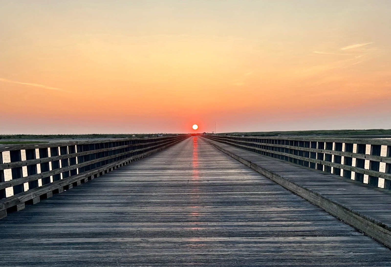 A wooden pier extending out over a marsh at sunset, dunes and water visible to either side of the horizon.